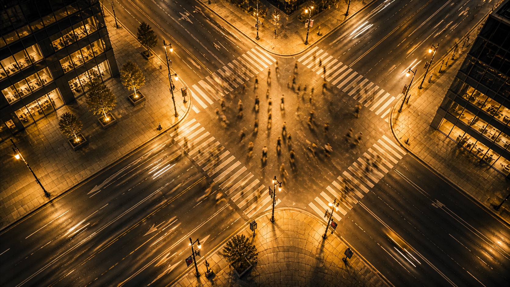 Aerial night view of a city intersection with pedestrians crossing in multiple directions, illuminated streetlights, and light trails from passing traffic.